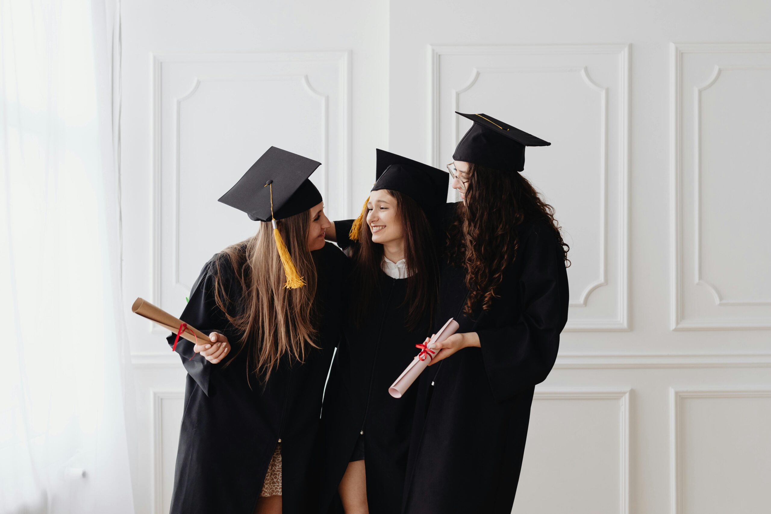 Three young women in graduation gowns and caps celebrate together indoors with diplomas in hand.